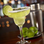 A margarita in a traditional glass on a dark wood surface with a bar in the background